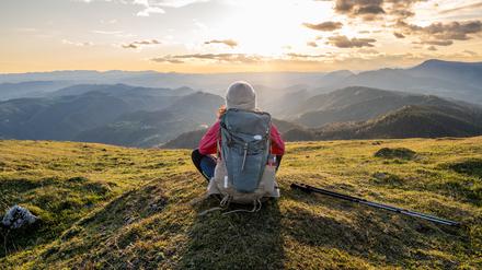 Eine Frau mit Mütze und Rucksack sitzt auf einem Berg und blickt über die Berglandschaft vor ihr. Sie ist nur von hinten zu sehen.