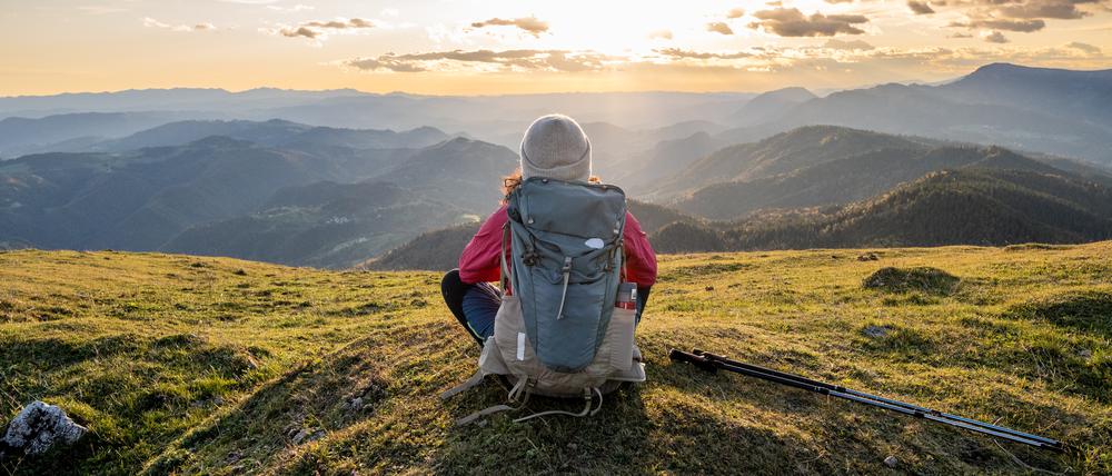 Eine Frau mit Mütze und Rucksack sitzt auf einem Berg und blickt über die Berglandschaft vor ihr. Sie ist nur von hinten zu sehen.