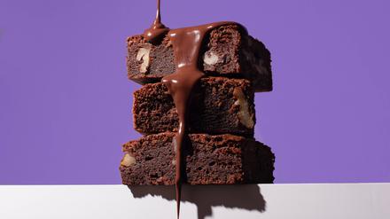 Stack of homemade chocolate brownies, each richly filled with nuts, being generously drizzled with a glossy chocolate sauce against a striking purple background. Model Released OlgaVila_ID23079_670986_003 Copyright: xOlgaxVilax