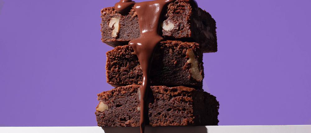 Stack of homemade chocolate brownies, each richly filled with nuts, being generously drizzled with a glossy chocolate sauce against a striking purple background. Model Released OlgaVila_ID23079_670986_003 Copyright: xOlgaxVilax