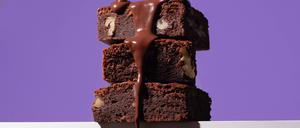 Stack of homemade chocolate brownies, each richly filled with nuts, being generously drizzled with a glossy chocolate sauce against a striking purple background. Model Released OlgaVila_ID23079_670986_003 Copyright: xOlgaxVilax