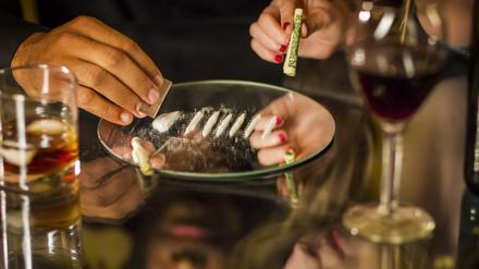 Woman's hand holding rolled dollar with man cutting cocaine line on table. Horizontal shot.