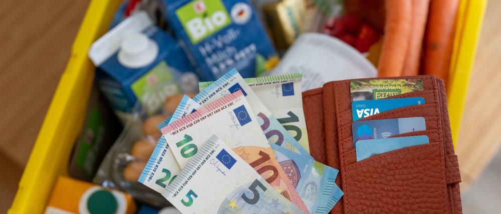 Eine Frau hält ein Portemonnaie mit Bargeld über einer Einkaufskiste mit Lebensmitteln. 