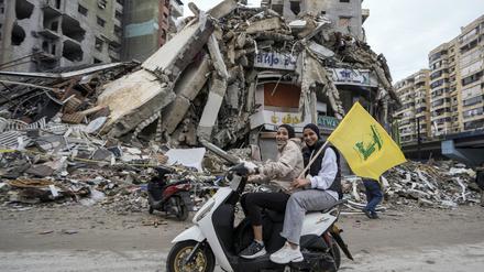 Frauen auf einem Motorroller halten eine Hisbollah-Fahne, während sie an einem zerstörten Gebäude vorbeifahren.