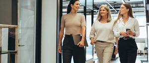 Drei Business-Frauen laufen beschwingt durch einen Flur. 