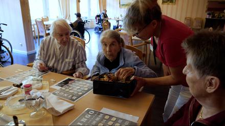 Bingo-Runde im Franz-Jordan-Stift in Berlin-Reinickendorf. Drei Frauen sitzen am Tisch mit den Bingo-Karten. Eine von ihnen nimmt sich ein Tütchen Gummibären.