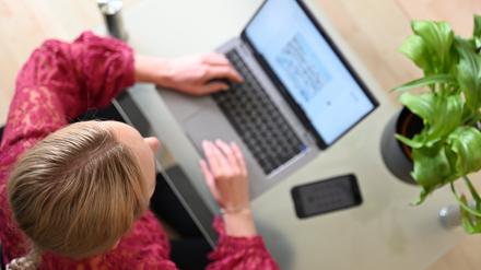 Eine Frau sitzt zu Hause an einem Glastisch und tippt auf einem Laptop, neben ihr liegt ein Smartphone.