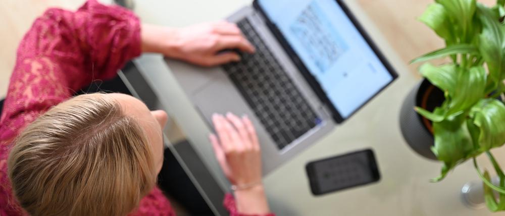 Eine Frau sitzt zu Hause an einem Glastisch und tippt auf einem Laptop, neben ihr liegt ein Smartphone.