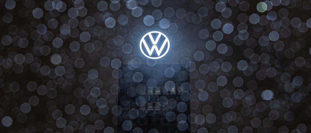 Wolfsburg: Regentropfen sammeln sich am frühen Morgen auf der Scheibe eines Autos, während dahinter das Logo auf dem Dach des VW-Markenhochhauses leuchtet.