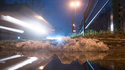 Autos fahren hinter einem kleinen Rest Schnee vorbei, der sich in einer Pfütze spiegelt.