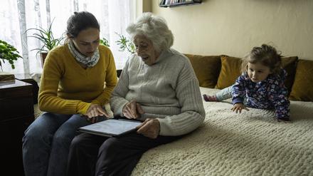 Eine Frau sitzt mit ihrer Mutter und ihrer Tochter auf einem Bett. Eine neue Studie zeigt, wie sehr die Doppelrolle von Elternpflege und Kindererziehung belasten kann.