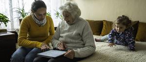 Eine Frau sitzt mit ihrer Mutter und ihrer Tochter auf einem Bett. Eine neue Studie zeigt, wie sehr die Doppelrolle von Elternpflege und Kindererziehung belasten kann.