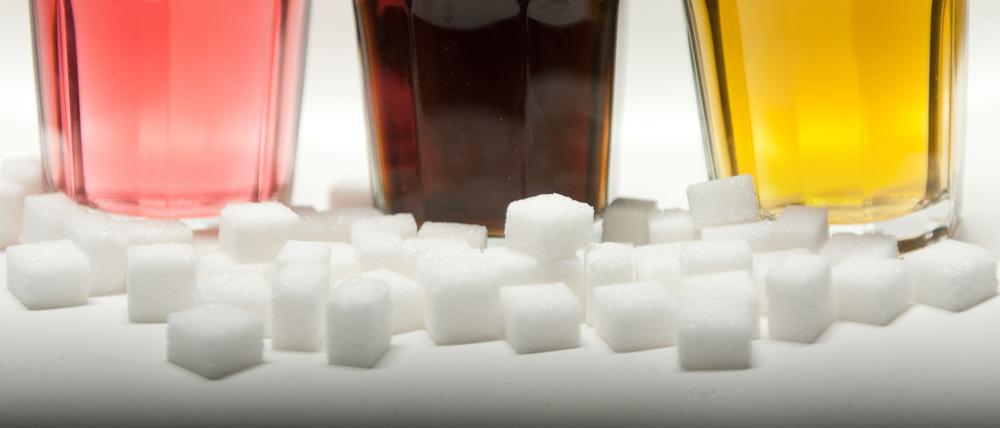 Drei Gläser gefüllt mit roter Limonade, Cola und einem Energy-Drink neben Zuckerwürfeln auf einem Tisch