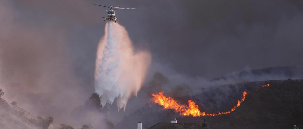 Ein Helikopter wirft am 7. Januar Wasser auf vom Palisades Feuer bedrohte Häuser in Los Angeles.  Nachlassende Winde könnten die Lage in den Waldbrandgebieten bald deutlich verbessern. 