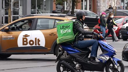 Ein Uber Eats-Kurier auf dem Roller und ein Auto mit Bolt-Logo im Hintergrund.