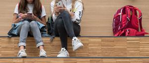 Schülerinnen sitzen im Foyer eines Gymnasium auf einer Freitreppe und halten jeweils ein Smartphone. 