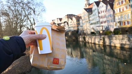 Eine McDonald’s To-Go-Tüte und ein To-Go-Becher werden auf der Neckarbrücke in Tübingen vor der Kulisse der Altstadt gehalten (gestellte Szene).