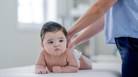 Ein Baby, das nur eine Windel trägt, liegt auf dem Bauch auf einer Behandlungsliege. Ein Erwachsener legt beide Hände auf den Rücken des Kindes.