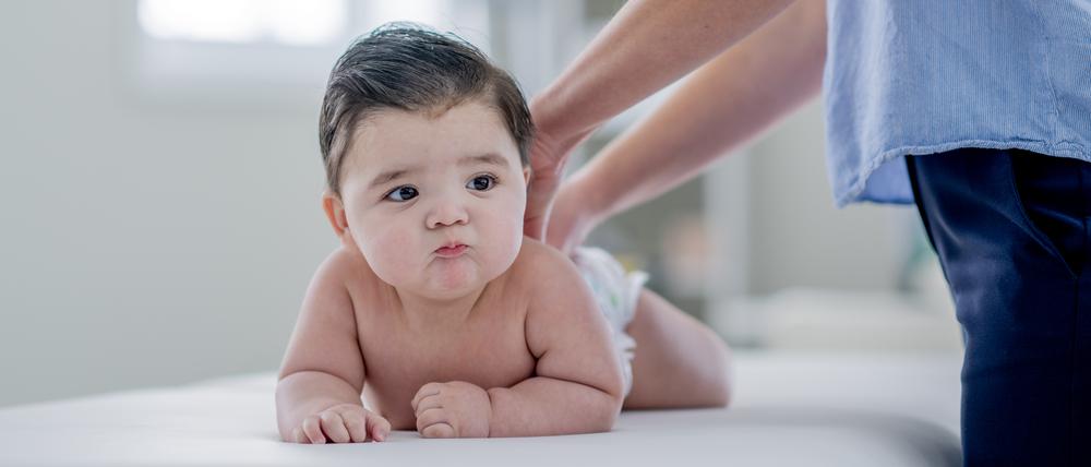 Ein Baby, das nur eine Windel trägt, liegt auf dem Bauch auf einer Behandlungsliege. Ein Erwachsener legt beide Hände auf den Rücken des Kindes.