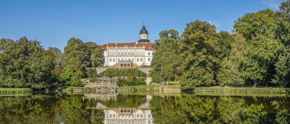 Blick auf das von Bäumen umgebene Schloss Wiesenburg und den davor liegenden See im Schlosspark.