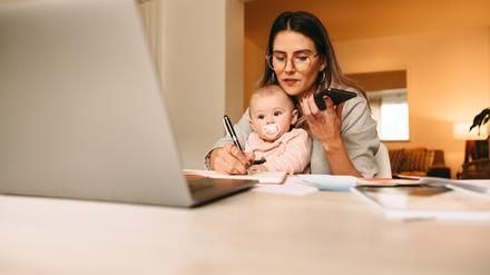 Eine Frau arbeitet vorm Laptop, Handy in der Hand, einen Stift in der anderen, das Kind auf dem Schoß. Zu wenig Kitas und verfehlte Gleichstellung hindern viele Mütter daran, vollzeit in den Job zurückzukehren.