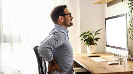 Ein Mann sitzt an seinem Schreibtisch und hält sich den schmerzenden Rücken.