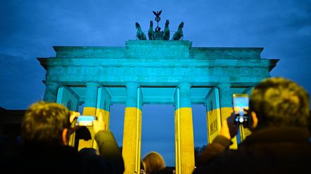 Passanten fotografieren vor einer Gedenkveranstaltung zum dritten Jahrestag des russischen Angriffs auf die Ukraine das Brandenburger Tor, das in den ukrainischen Nationalfarben angestrahlt ist.