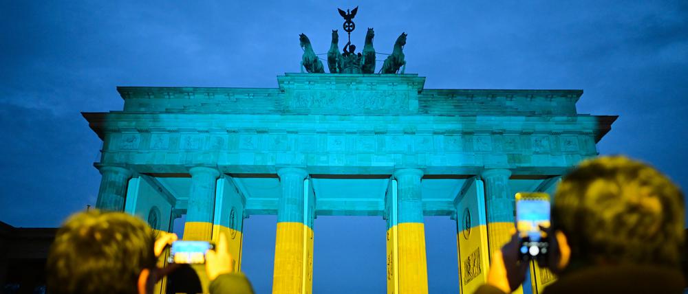 Passanten fotografieren vor einer Gedenkveranstaltung zum dritten Jahrestag des russischen Angriffs auf die Ukraine das Brandenburger Tor, das in den ukrainischen Nationalfarben angestrahlt ist.