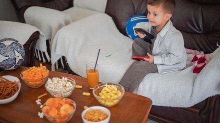 Ein etwa drei- bis vierjähriges Kind sitzt mit einer Fernbedienung auf einem Sofa. Auf dem Tisch vor ihm stehen mehrere Schüsseln mit Chips und ein Glas Saft.