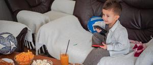 Ein etwa drei- bis vierjähriges Kind sitzt mit einer Fernbedienung auf einem Sofa. Auf dem Tisch vor ihm stehen mehrere Schüsseln mit Chips und ein Glas Saft.
