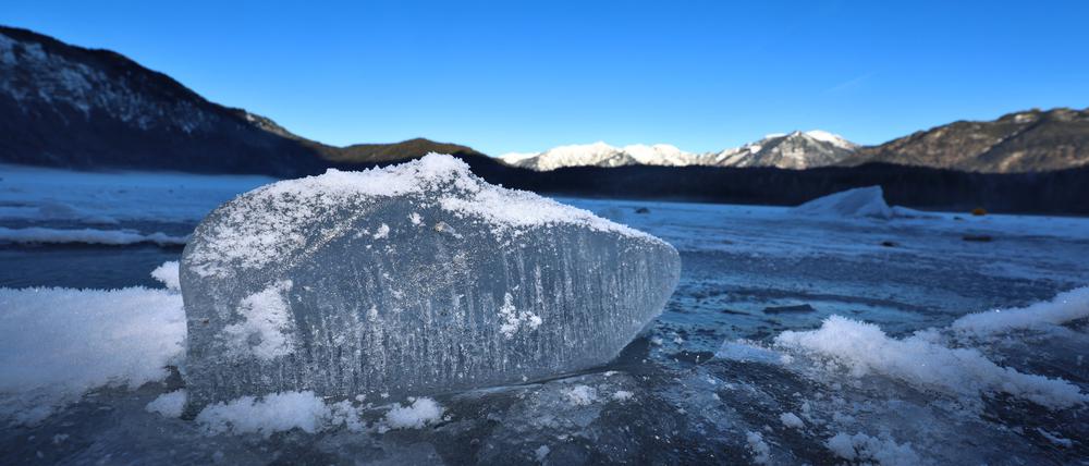 Ein Eisbrocken liegt auf dem mit einer Eisdecke überzogenen Eibsee unter strahlend blauem Himmel.