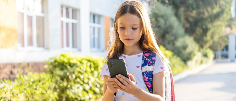 Ein Mädchen mit einem Schulranzen schaut auf ein Handy.