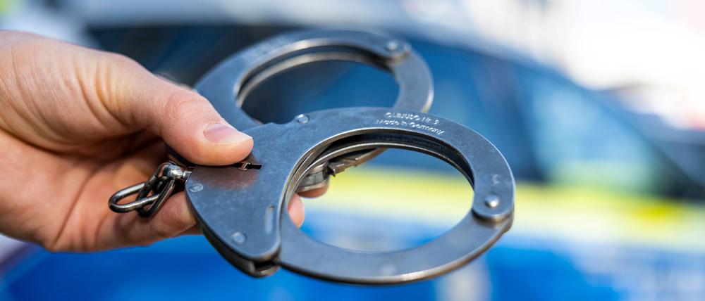 Eine Hand hält Handschellen vor einen Streifenwagen der Polizei. (Symbolbild)