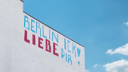 Ein blau-rotes Graffito mit der Aufschrift „Berlin Ick liebe Dir“ steht auf einer weißen Wand eines hohen Gebäudes, dahinter blauer Himmel und ein, zwei Wölkchen.