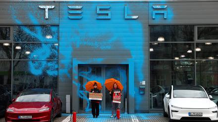 Vor der mit blauer Farbe besprühten Front des Tesla-Centers in Reinickendorf stehen zwei Frauen mit orangen Regenschirmen.    