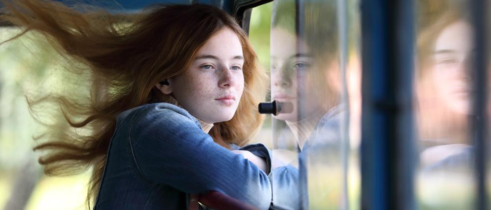 Eine junge Frau steht mit wehenden Haaren an einem offenen Fenster.