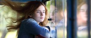 Eine junge Frau steht mit wehenden Haaren an einem offenen Fenster.