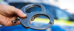 Eine Hand hält Handschellen vor einen Streifenwagen der Polizei. 
