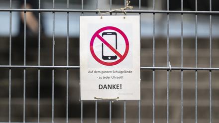 Ein Schild mit einem durchgestrichenen Smartphone und der Aufschrift «Auf dem ganzen Schulgelände - zu jeder Uhrzeit! DANKE!» hängt am Zugangstor zum Schulhof eines Gymnasiums und weist auf ein generelles Handyverbot hin. 