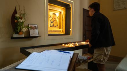 Ein Kondolenzbuch zum Tod von Papst Franziskus liegt in der Katholischen Probsteikirche St. Peter und Paul in Potsdam vor einem kleinen Foto des Papstes. Im Hintergrund hat ein junger Mann eine Kerze angezündet.