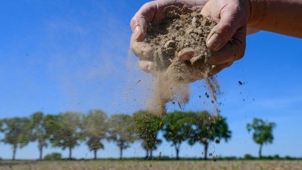 Eine Frau zeigt trockenen Ackerboden auf einem Feld in Brandenburg.