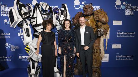 Sabiha Ghellal (l-r), Dorothee Bär (CSU), Bundesministerin für Forschung, Technologie und Raumfahrt, und Felix Falk stehen am Rand der Verleihung des Deutschen Computerspielpreises im Palais am Funkturm vor Figuren von Computerspielcharakteren. 