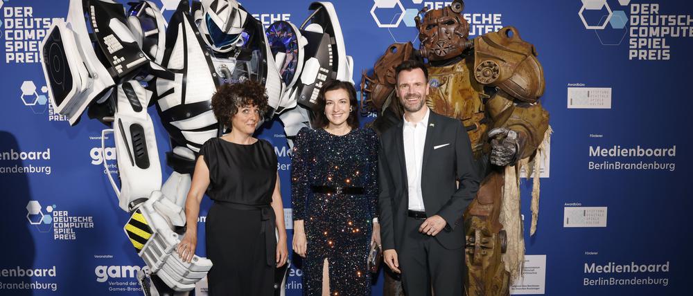 Sabiha Ghellal (l-r), Dorothee Bär (CSU), Bundesministerin für Forschung, Technologie und Raumfahrt, und Felix Falk stehen am Rand der Verleihung des Deutschen Computerspielpreises im Palais am Funkturm vor Figuren von Computerspielcharakteren.
