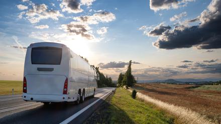 Ein weißer Reisebus fährt entlang eines Feldes auf einen Sonnenuntergang zu.