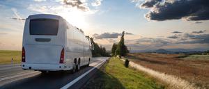 Ein weißer Reisebus fährt entlang eines Feldes auf einen Sonnenuntergang zu.