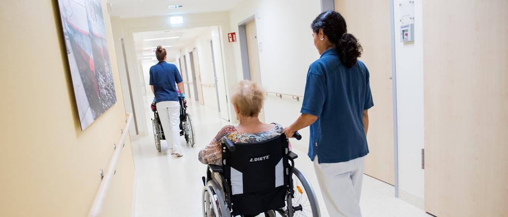 Zwei Pflegerinnen schieben ältere Patienten mit Rollstühlen durch einen Flur.