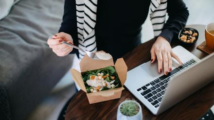 Eine Frau isst, während sie an einem Laptop sitzt und arbeitet.