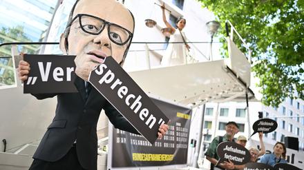Ein Aktivist mit Merz-Maske zerbricht während einer symbolischen Protestaktion von Kabul Luftbrücke und Campact ein Schild mit der Aufschrift „Versprechen“. 