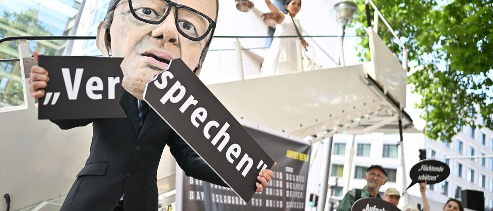Ein Aktivist mit Merz-Maske zerbricht während einer symbolischen Protestaktion von Kabul Luftbrücke und Campact ein Schild mit der Aufschrift „Versprechen“.
