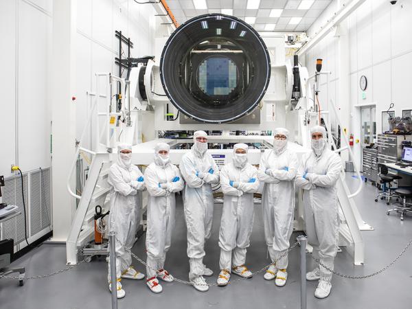 A group of six people stand in front of the huge LSST digital camera, which is oriented so that we're looking into the lens opening and at the blue-tinted tiled focal plane of CCDs. The group of people are all outfitted head to toe in white clean room garb, with hair caps, face masks, white onesies, and foot covers. Credit: Jacqueline Ramseyer Orrell/SLAC National Accelerator Laboratory